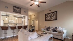 A living room with a sofa, a coffee table, and a ceiling fan. There is a kitchen area with a microwave and bar stools. - FAMILY RETREAT/ PET FRIENDLY NEAR MILITARY BEACHES AND BASES (Jacksonville)