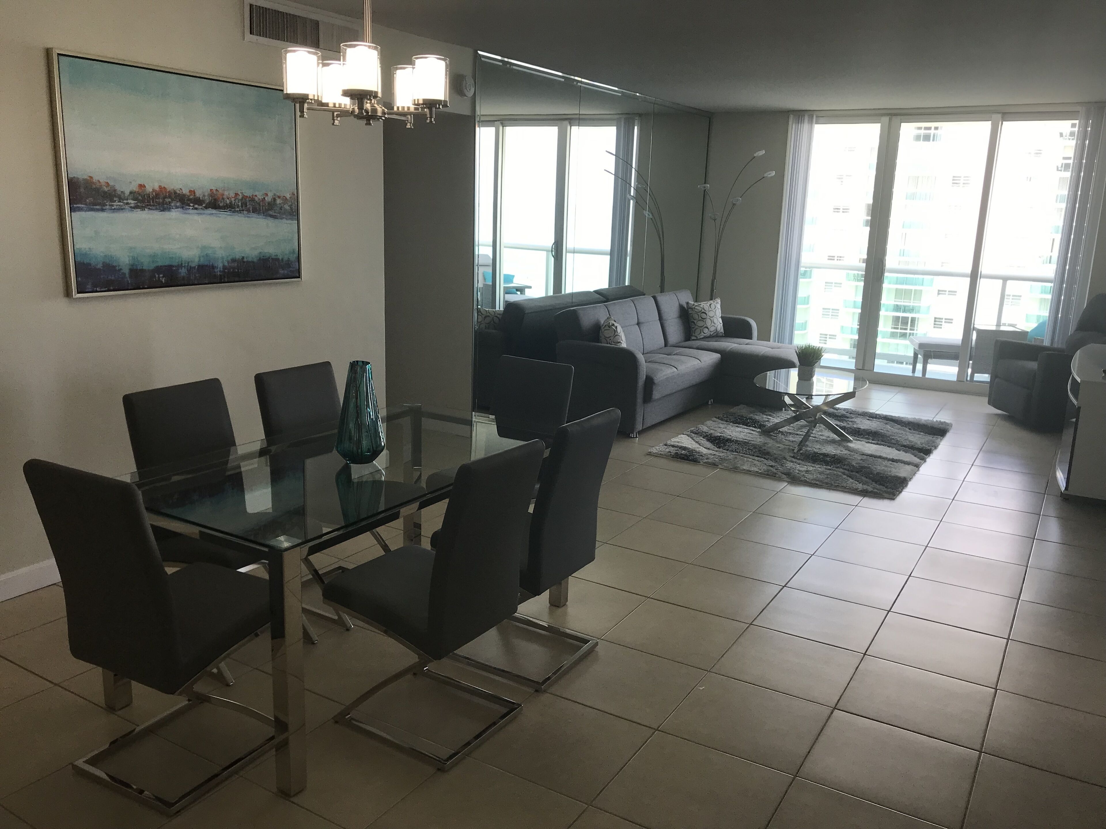 A modern living room with a glass dining table, grey sofa, and a large window offering a view of buildings.