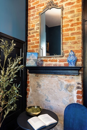 A room with a brick wall, a mirror, a book, a bowl, and a blue chair.