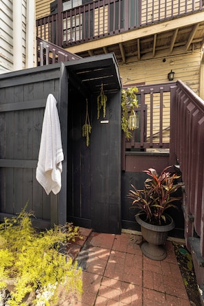 A wooden outdoor shower area with a towel hanging on the door, surrounded by plants and a potted plant.