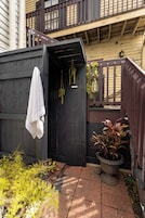 A wooden outdoor shower area with a towel hanging on the door, surrounded by plants and a potted plant.