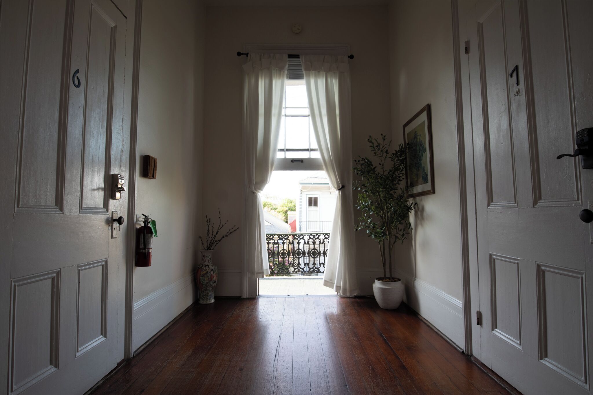 A hallway with wooden flooring, white doors, and a view to the outside through a window with white curtains.