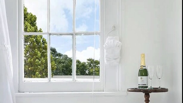 A bathroom with a freestanding bathtub, a window with a view of greenery, and a small table with a bottle and glasses. - The Mansion on Royal (New Orleans)