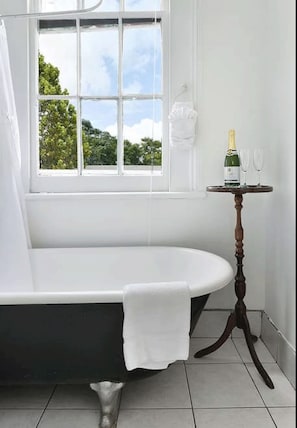 A bathroom with a freestanding bathtub, a window with a view of greenery, and a small table with a bottle and glasses. - The Mansion on Royal (New Orleans)