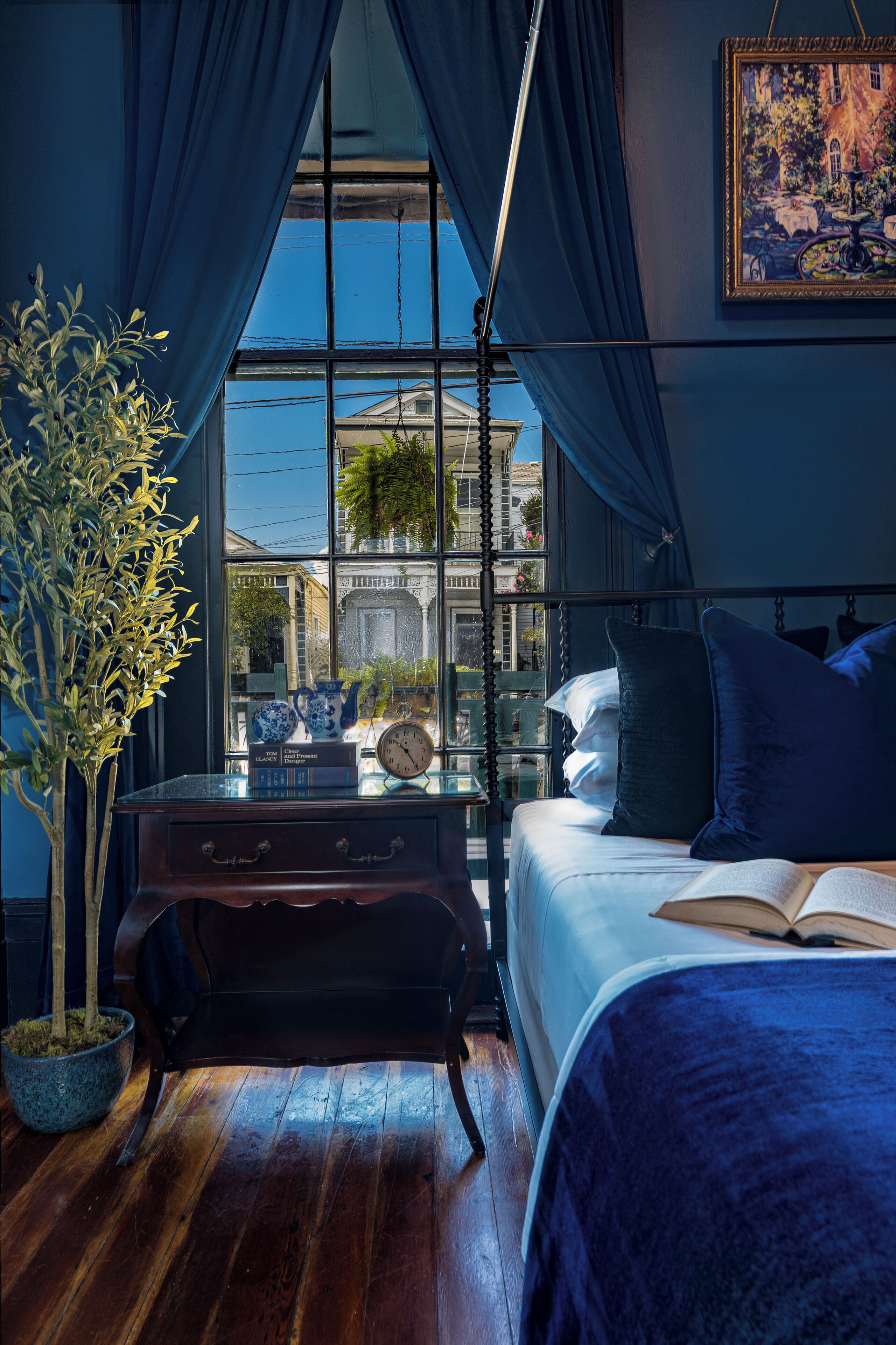 A bedroom with a large window, a bed, a bedside table with books and a clock, and a potted plant.