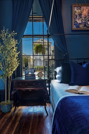 A bedroom with a large window, a bed, a bedside table with books and a clock, and a potted plant. - The Mansion on Royal (New Orleans)