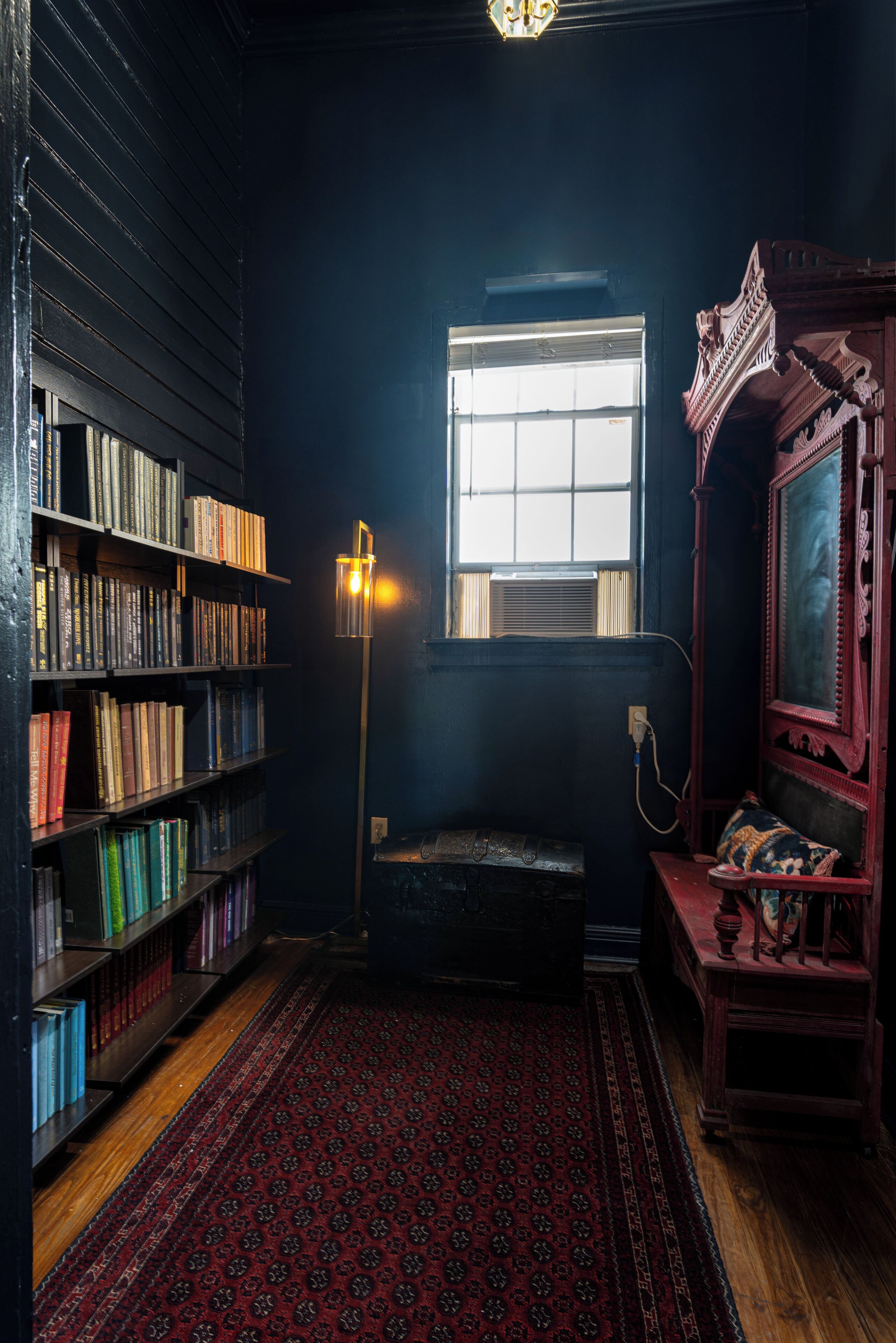 Une pièce avec une bibliothèque, une lampe, une fenêtre, un miroir et un tapis à motifs.