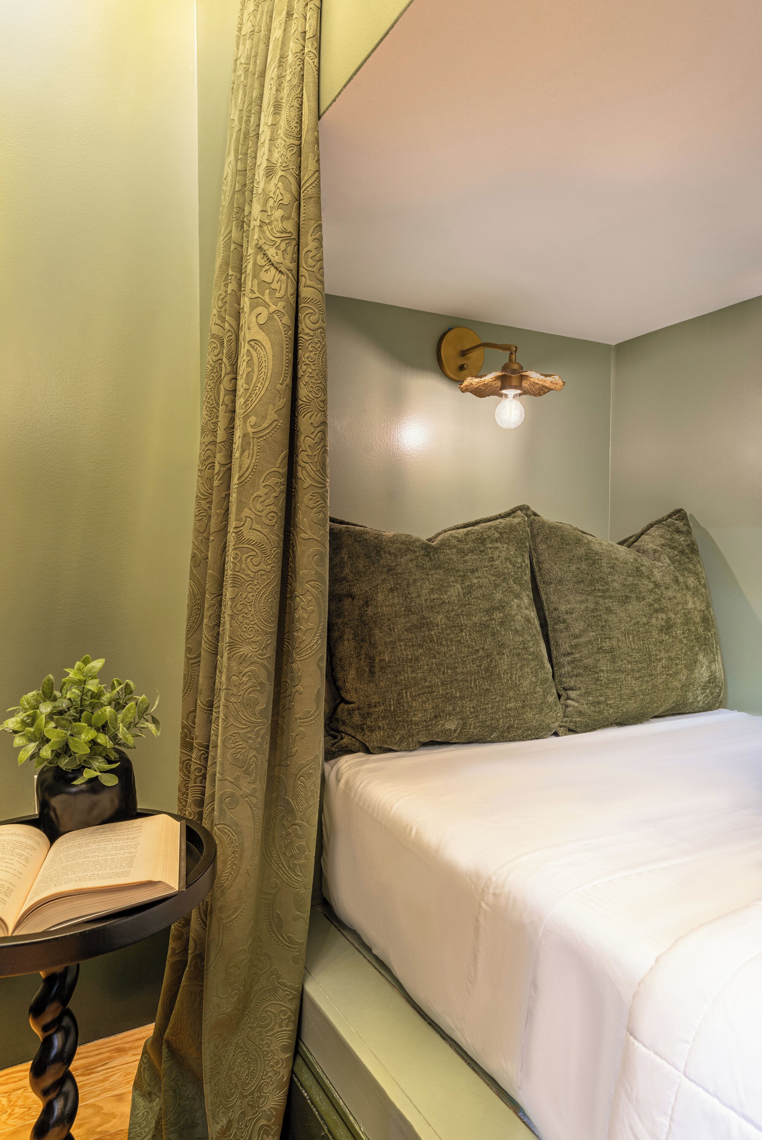 A room with a bed, a bedside table with a book and a plant, and a curtain.