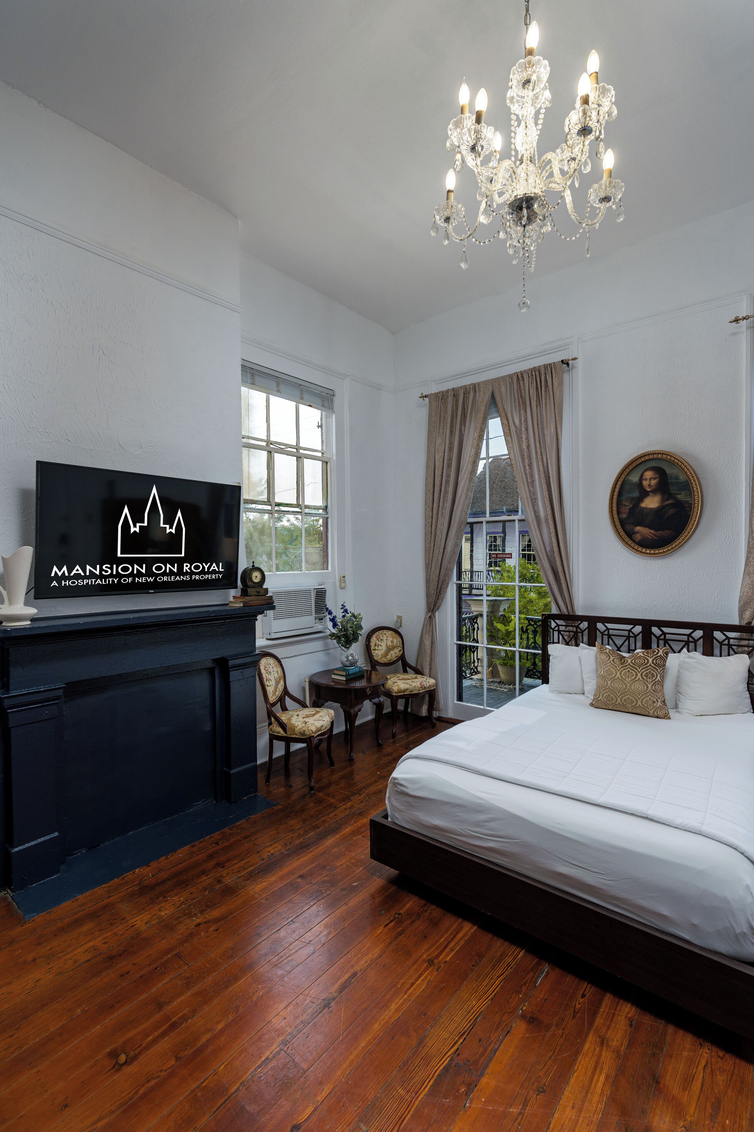 A hotel room with a bed, a television, a chandelier, a painting, and a view of the outdoors.