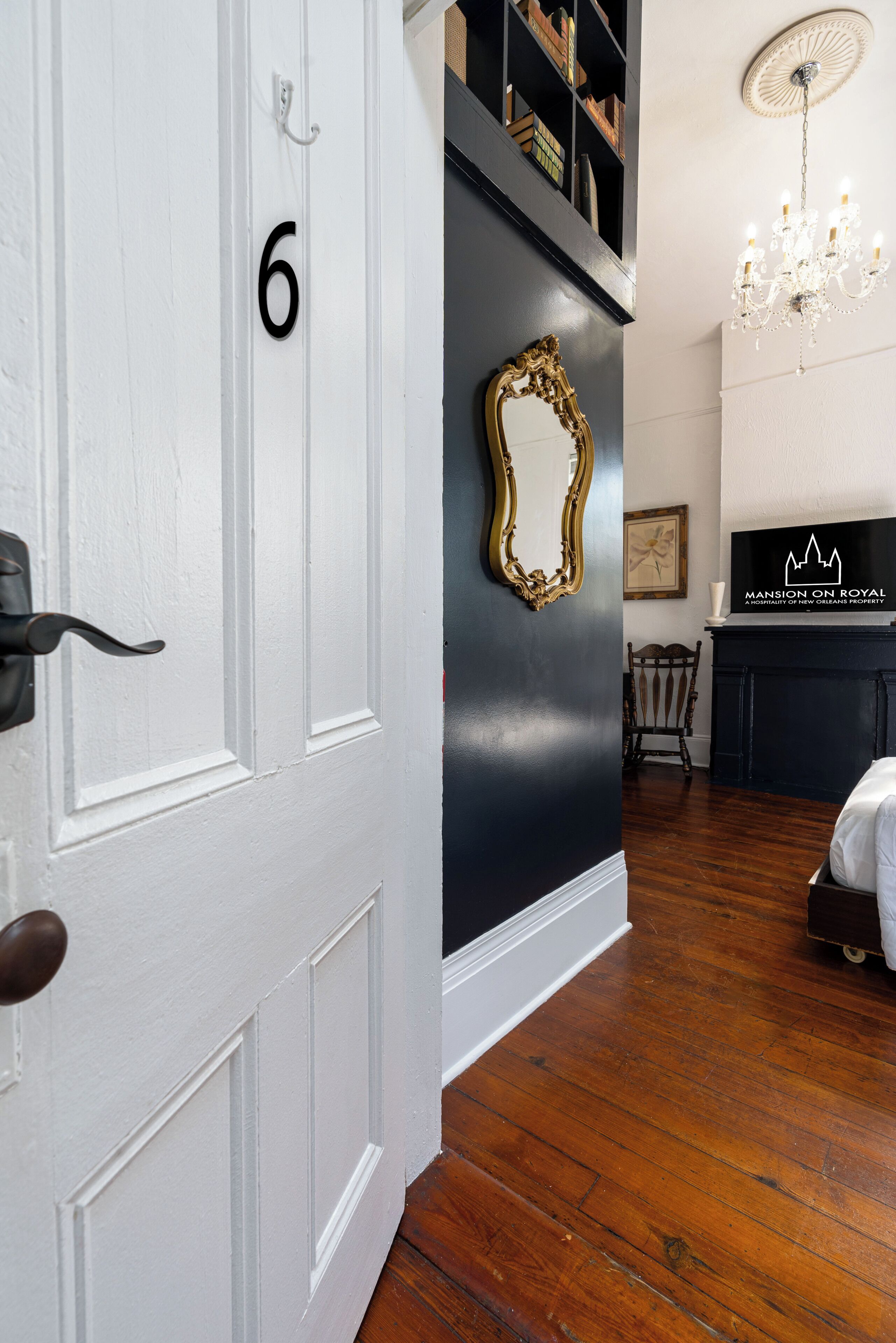 Une pièce avec une porte blanche, un mur noir orné d’un miroir doré, un lustre et une enseigne indiquant « Mansion on Royal ».
