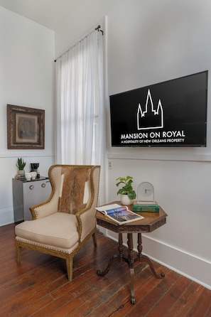 A room with a wooden chair, a small table with a book and a plant, a framed picture on the wall, and a TV displaying 'MANSION ON ROYAL'. - The Mansion on Royal (New Orleans)