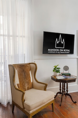A room with a large window, a chair with a fur throw, a small table with a plant and books, and a wall-mounted TV displaying 'MANSION ON ROYAL'. - The Mansion on Royal (New Orleans)