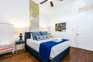Un dormitorio con cama, mesitas de noche, ventilador de techo, una puerta y un cuadro en la pared.