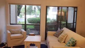 Ein Wohnzimmer mit zwei Sofas, einem kleinen Tisch und großen Fenstern mit Blick auf einen Garten und eine Straße.