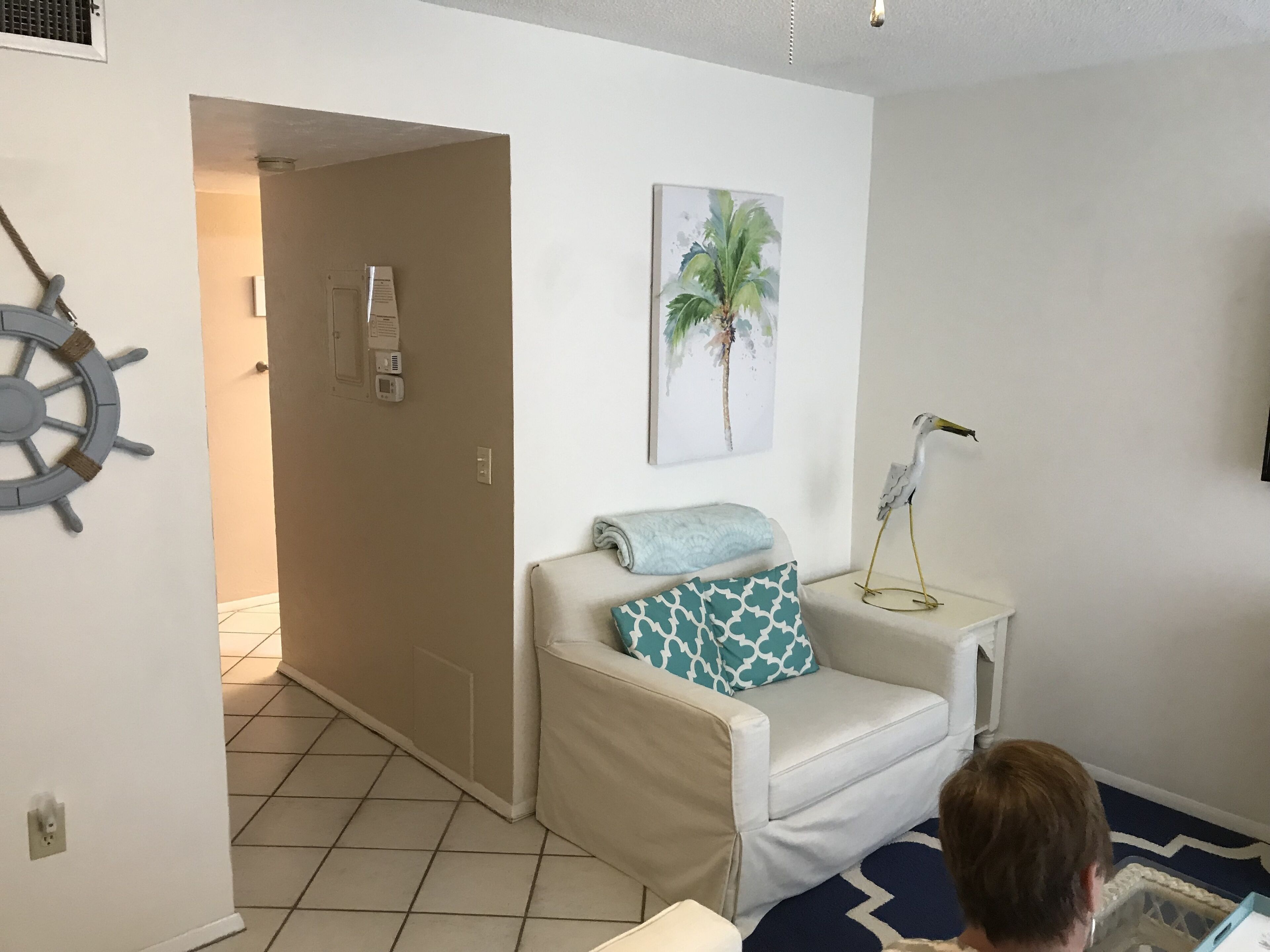 A room with a white sofa, a small table with a lamp, a wall-mounted painting of a palm tree, and a ship's wheel decoration.