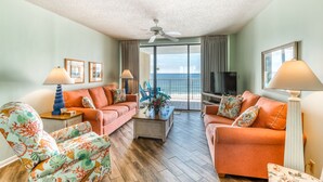 A living room with two orange sofas, a coffee table, and a TV. There are two lamps, a ceiling fan, and a large window with a view of the ocean. - Summer House on Romar Beach (Orange Beach)
