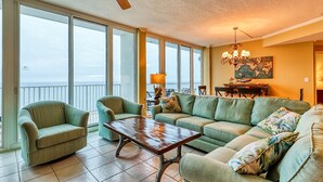 A spacious living room with a view of the ocean, a ceiling fan, and a chandelier. - Lighthouse Condominiums (Gulf Shores)