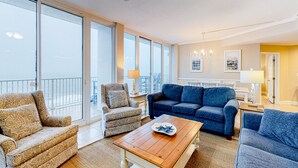 A spacious living room with a large window, a blue sofa, a patterned armchair, and a wooden coffee table. - Lighthouse Condominiums (Gulf Shores)