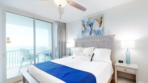 A hotel room with a large bed, a nightstand with a lamp, a painting on the wall, and a balcony with chairs. - Lighthouse Condominiums (Gulf Shores)