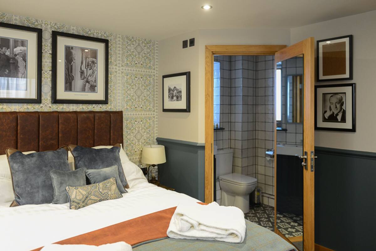 Habitación de hotel con cama, almohadas, mesita de noche con lámpara, baño con inodoro y lavabo, y cuadros en las paredes.