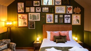 Quarto com uma cama grande, várias gravuras de plantas emolduradas na parede, um abajur de chão e uma poltrona com padrão floral.