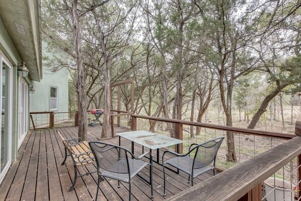 A wooden deck with outdoor furniture, including a table and chairs, overlooking a forested area. - Hill Country River House - Blanco River - Wilson Creek (Wimberley)