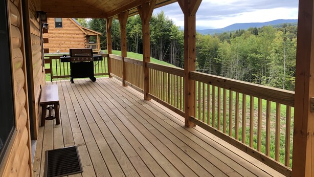 Eine Holzterrasse mit Grill und Bank, mit Blick auf eine bewaldete Landschaft und Berge im Hintergrund.