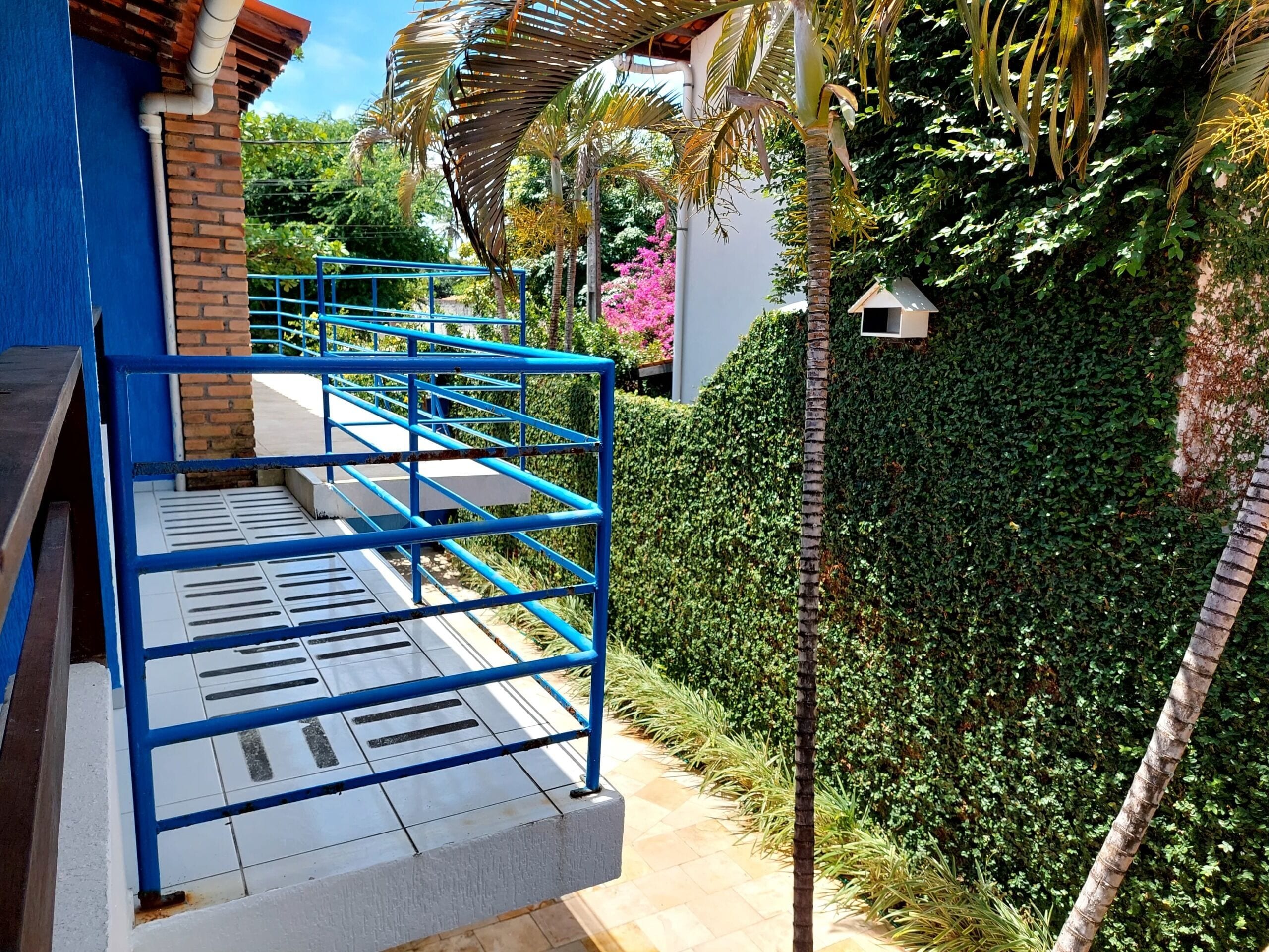 Una escalera azul conduce a un balcón con vistas a un jardín exuberante y un edificio de color azul.