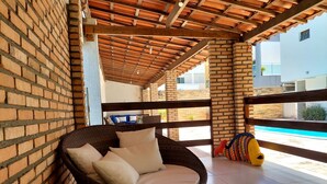 A wicker chair with cushions, a tiled floor, and a view of a swimming pool. - Pousada Lua Azul (Ipojuca)