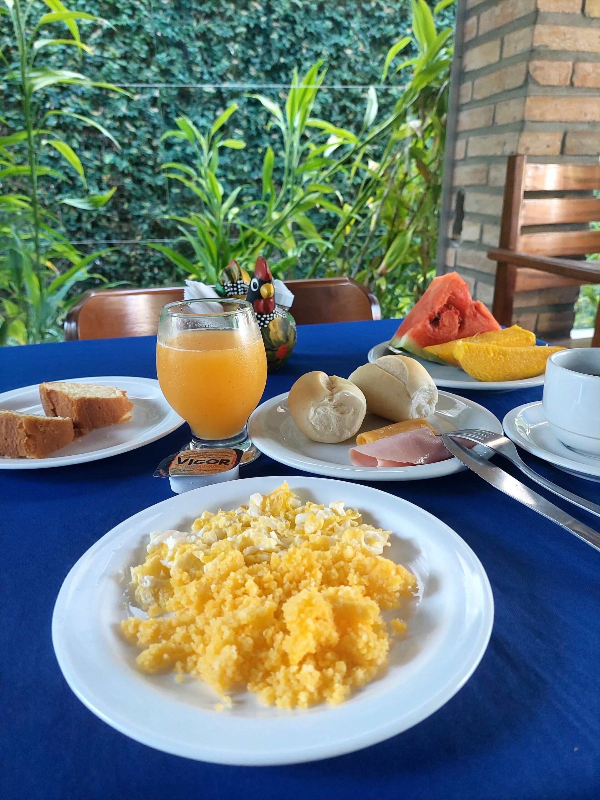 Una mesa de desayuno con platos de huevos revueltos, tostadas, jamón y fruta, junto con un vaso de jugo de naranja.