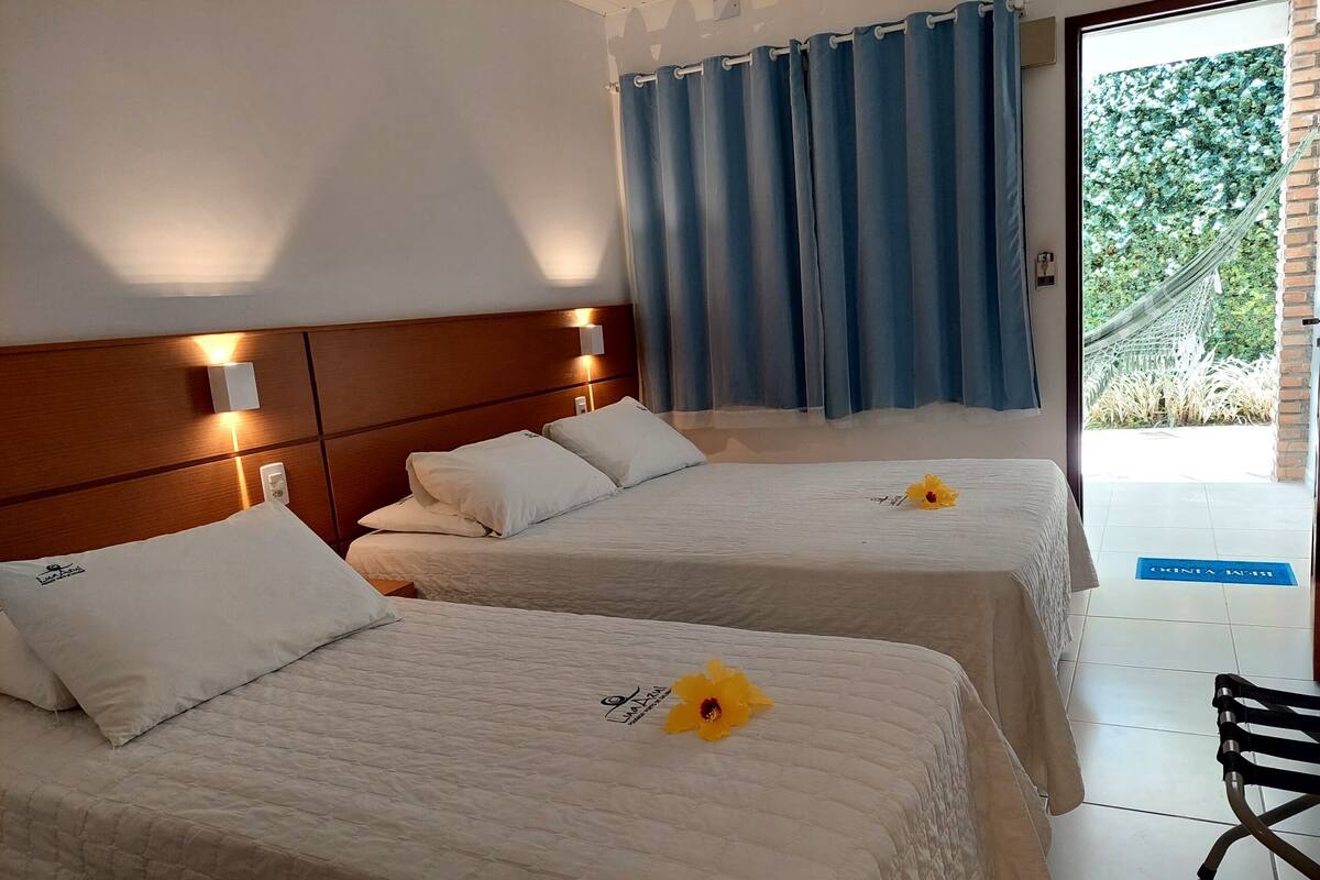 A hotel room with two beds, a wooden headboard, a window with blue curtains, and a view of greenery outside.
