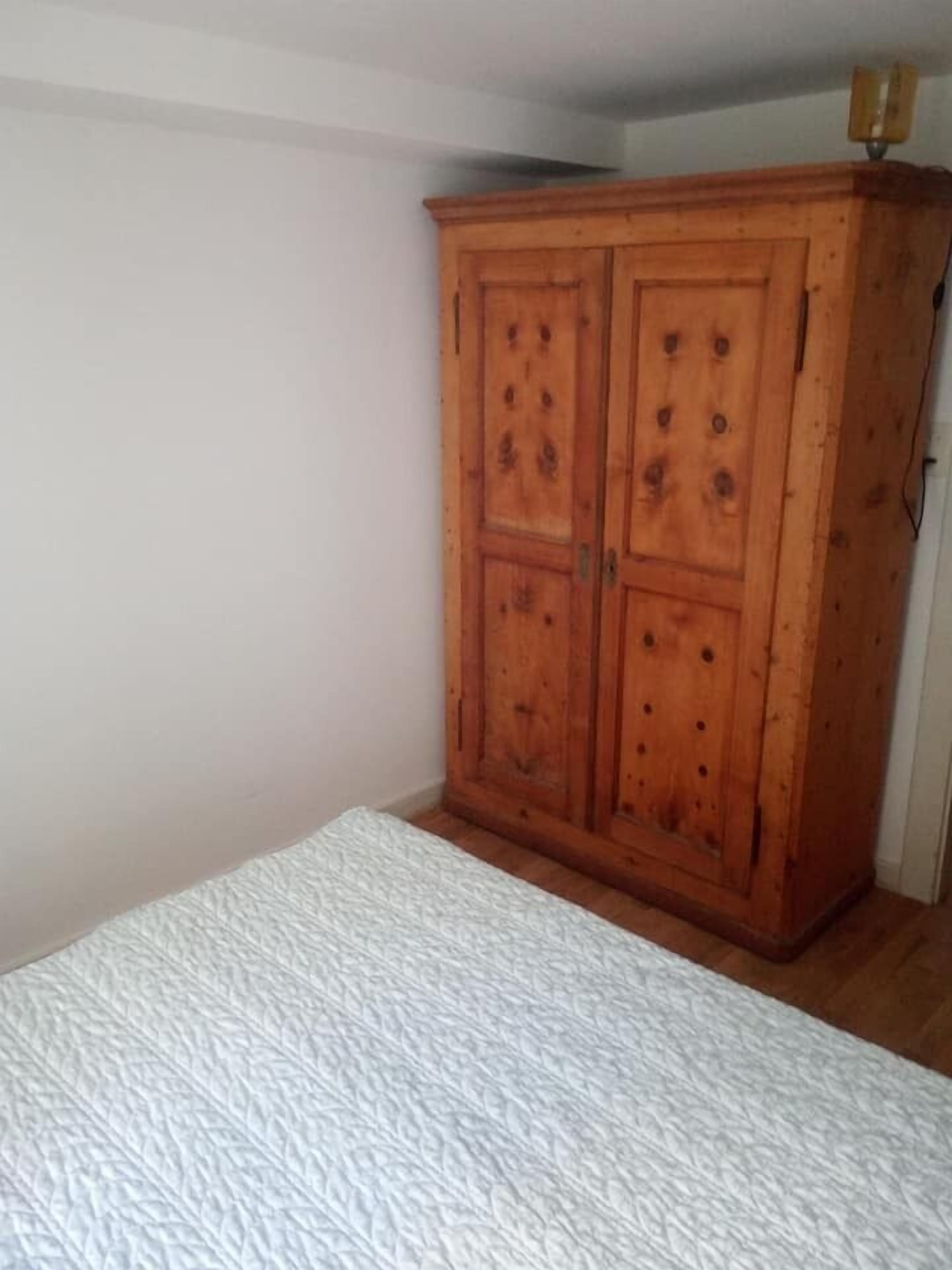 A bedroom with a wooden wardrobe, a white textured bedspread, and a plain white wall.