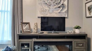Een woonkamer met een flatscreen televisie, een groot abstract schilderij en een entertainmentkast met glazen deuren, versierd met decoratieve objecten.