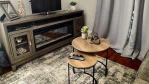 Een woonkamer met een donker houten entertainmentkast, een flatscreen televisie, twee ronde houten bijzettafeltjes, een afstandsbediening, een potplant en een ingelijste foto.