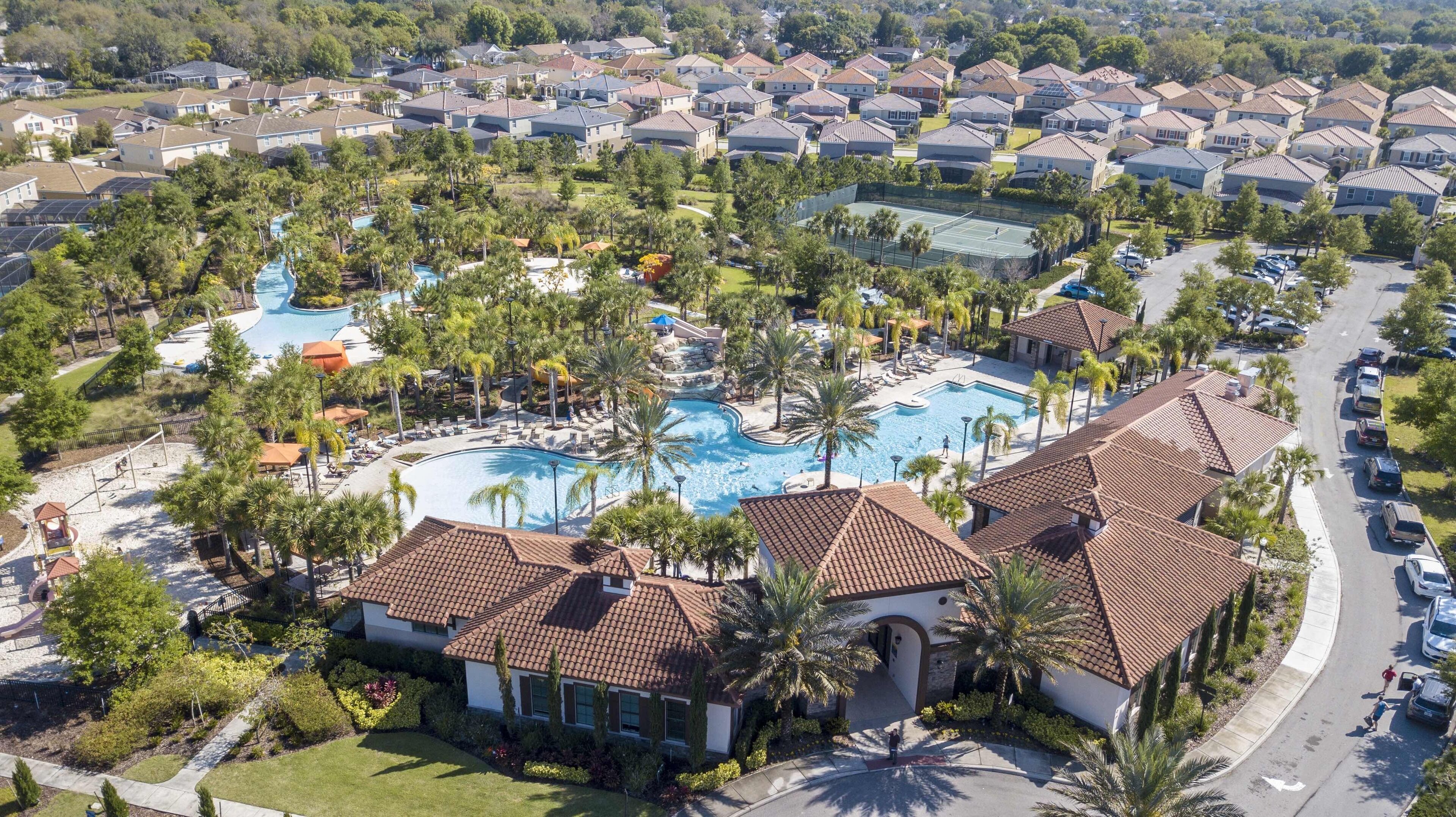A residential area with a large swimming pool surrounded by palm trees and houses with red-tiled roofs.