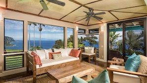 A spacious room with a large window offering a view of the ocean and palm trees. There is a wooden sofa, a coffee table, and a chair with a view. - Luxury 4BR Estate with Ocean Views, Pool, Slide & Gourmet Kitchen: Waipio Heaven (Honokaa)