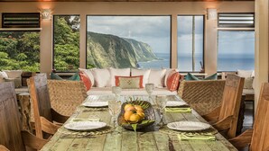 A dining area with a wooden table set for six, large windows with a coastal view, and a sofa with cushions. - Luxury 4BR Estate with Ocean Views, Pool, Slide & Gourmet Kitchen: Waipio Heaven (Honokaa)