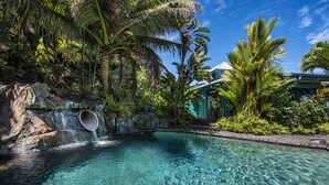 A clear pool with a waterfall feature, surrounded by lush tropical vegetation and a building with a green roof. - Luxury 4BR Estate with Ocean Views, Pool, Slide & Gourmet Kitchen: Waipio Heaven (Honokaa)