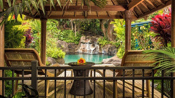 A wooden deck with a table and chairs overlooking a tropical garden with a waterfall and a pool. - Luxury 4BR Estate with Ocean Views, Pool, Slide & Gourmet Kitchen: Waipio Heaven (Honokaa)