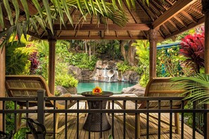 A wooden deck with a table and chairs overlooking a tropical garden with a waterfall and a pool.