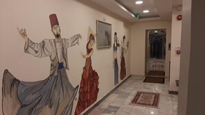 Un couloir orné d’une fresque représentant des danseurs, un tableau encadré accroché au mur et une porte portant un panneau indiquant la présence d’un ascenseur.