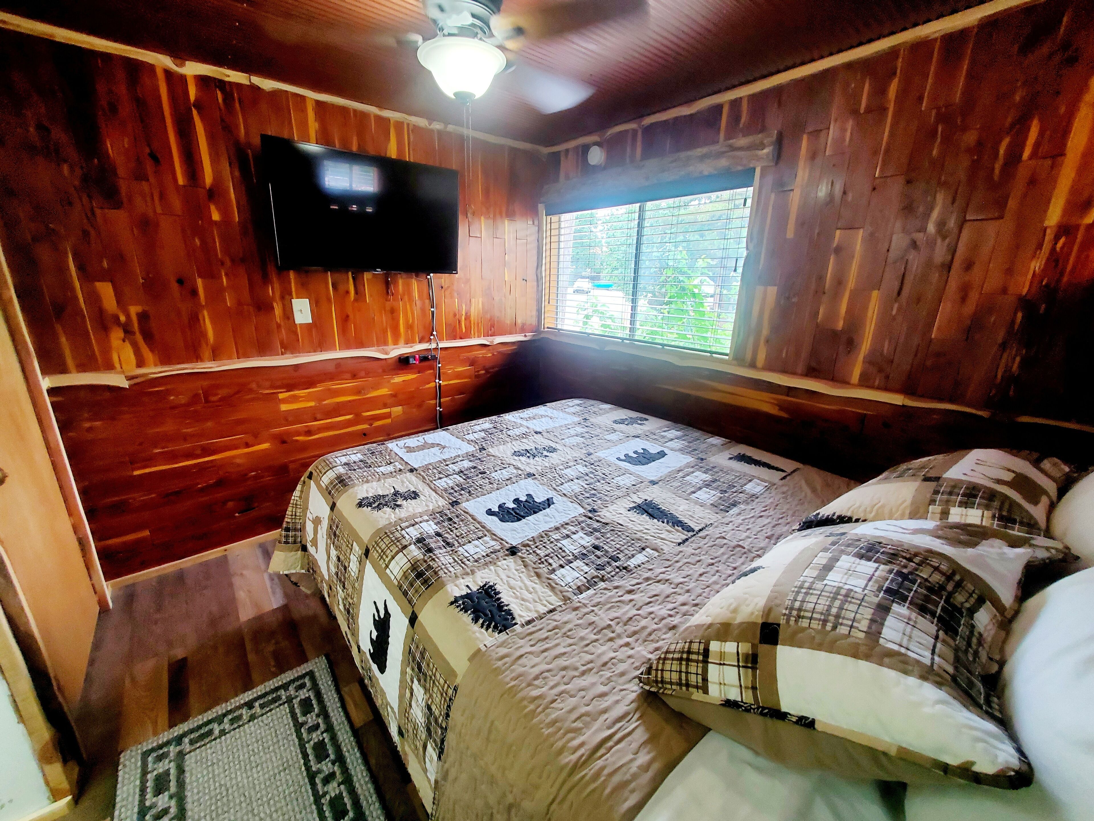 Habitación de cabaña de madera con una cama, un ventilador de techo, un televisor montado en la pared y una ventana con vistas a la vegetación.