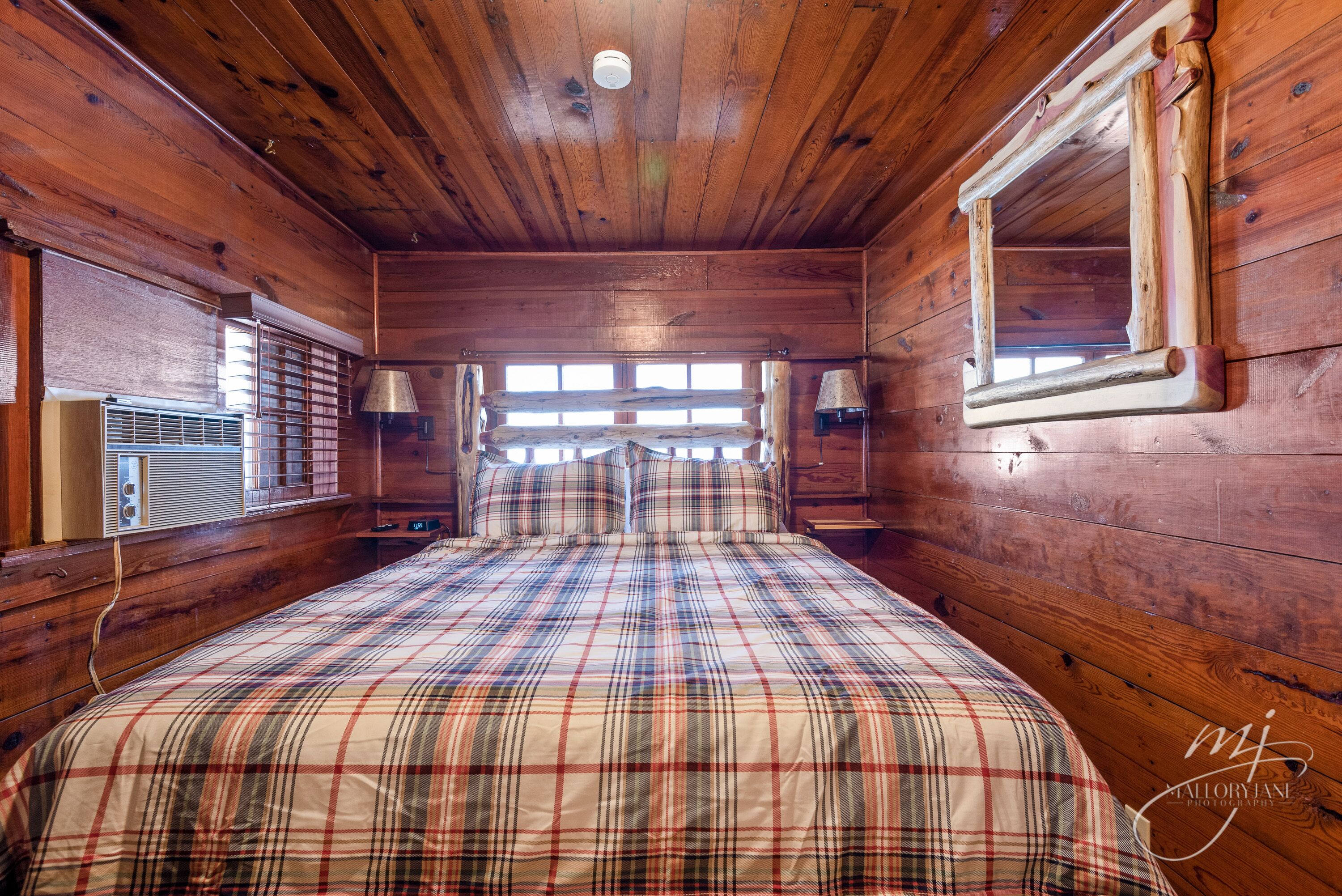 A wooden cabin room with a plaid bedspread, a window with blinds, and a wall-mounted lamp.