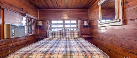 A wooden cabin room with a plaid bedspread, a window with blinds, and a wall-mounted lamp.