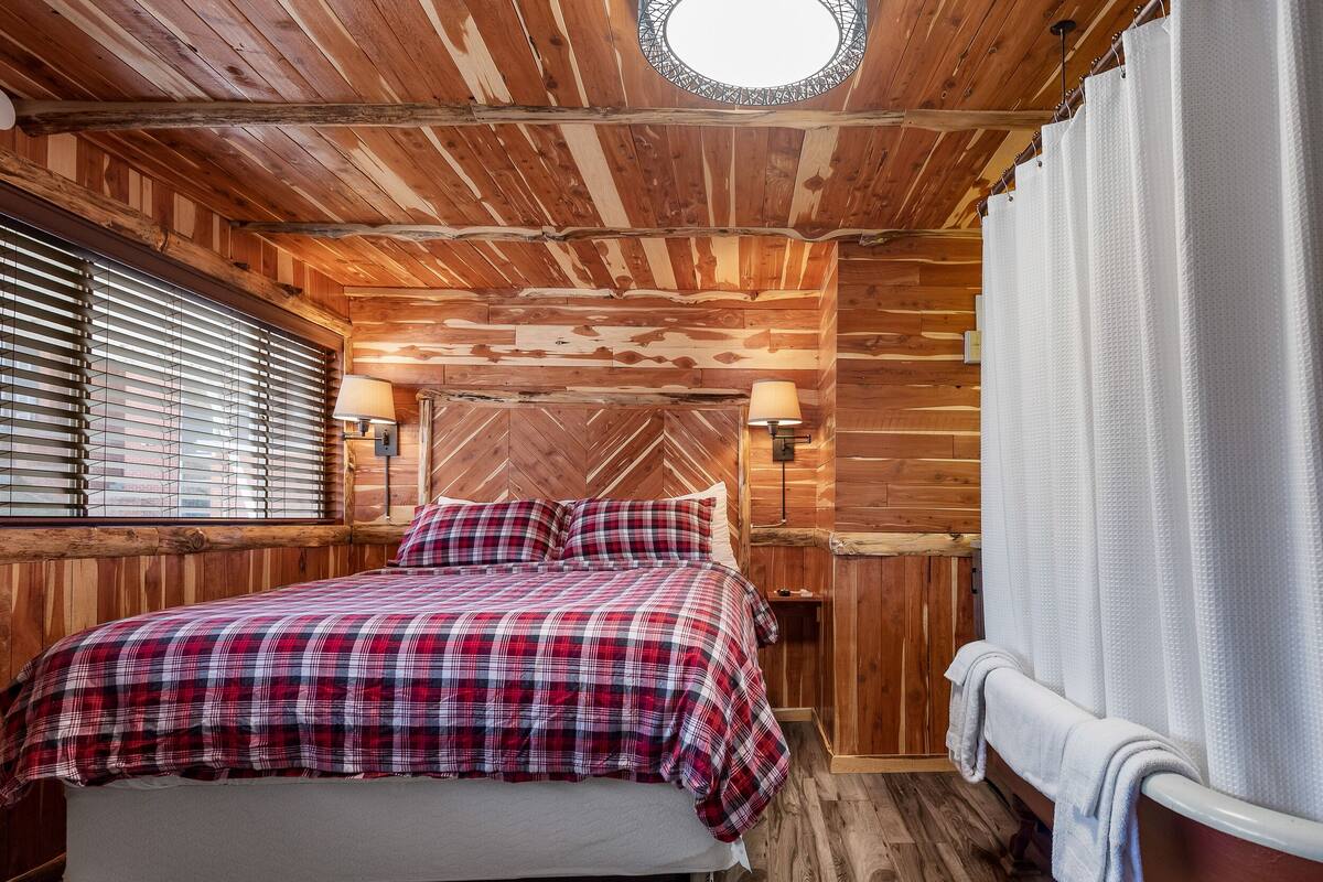 Un dormitorio de cabaña de madera con una cama, un ventanal con persianas, un tragaluz redondo y un baño con cortina de ducha.