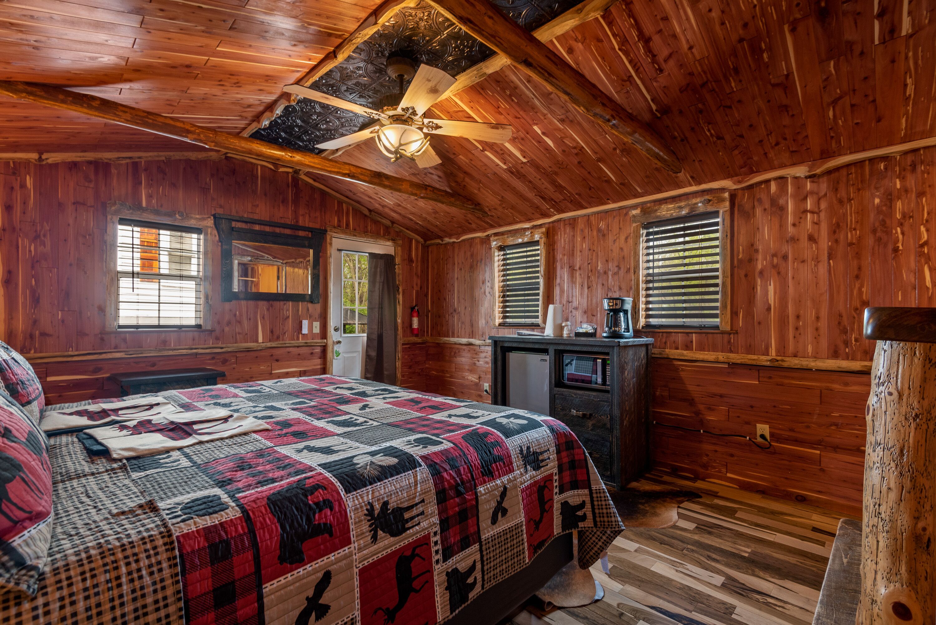 A wooden cabin bedroom with a large bed, a ceiling fan, and a small desk with a coffee maker.