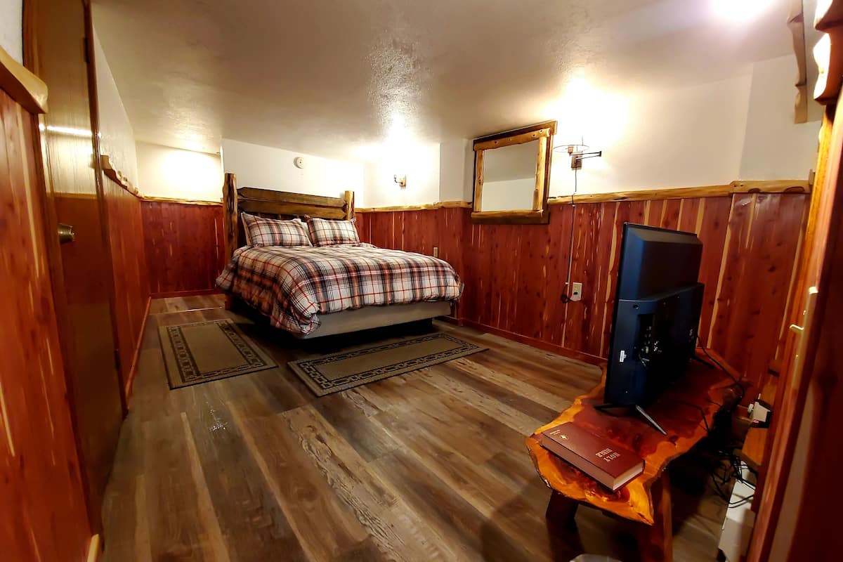 Un dormitorio con una cama de madera, un televisor sobre un mueble de madera y una pequeña mesa con un libro.