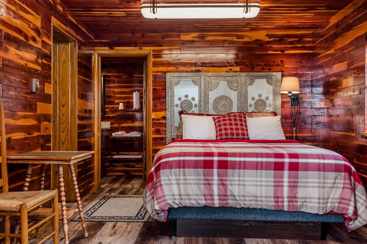 Un dormitorio de cabaña de madera con una cama, una silla, una mesa y una lámpara.