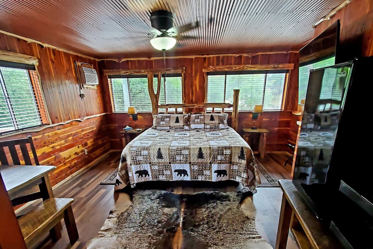 Una habitación de cabaña de troncos con una cama, bancos de madera, un tapiz de piel grande y ventanas con persianas.