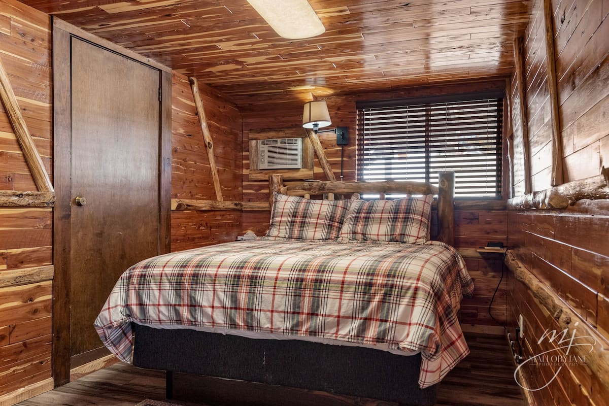 Un dormitorio de cabaña de madera con colcha a cuadros, una puerta de madera y una ventana con persianas.